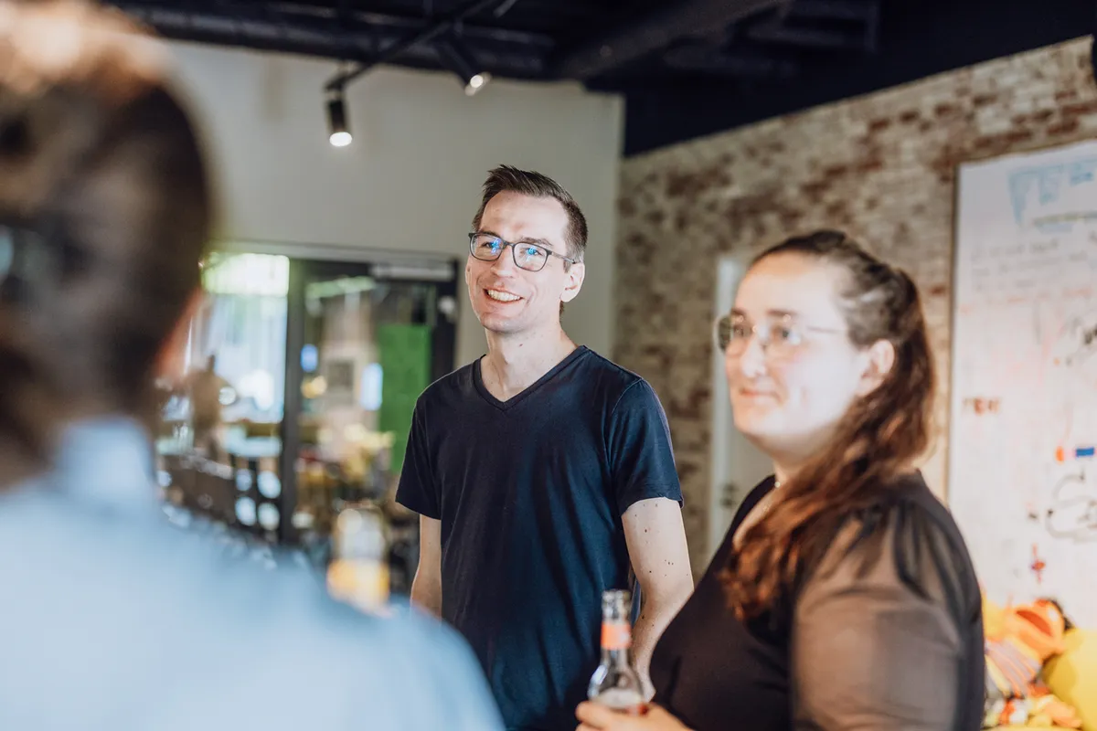 A group of people standing and smiling in a casual indoor setting, holding drinks and engaging in conversation.