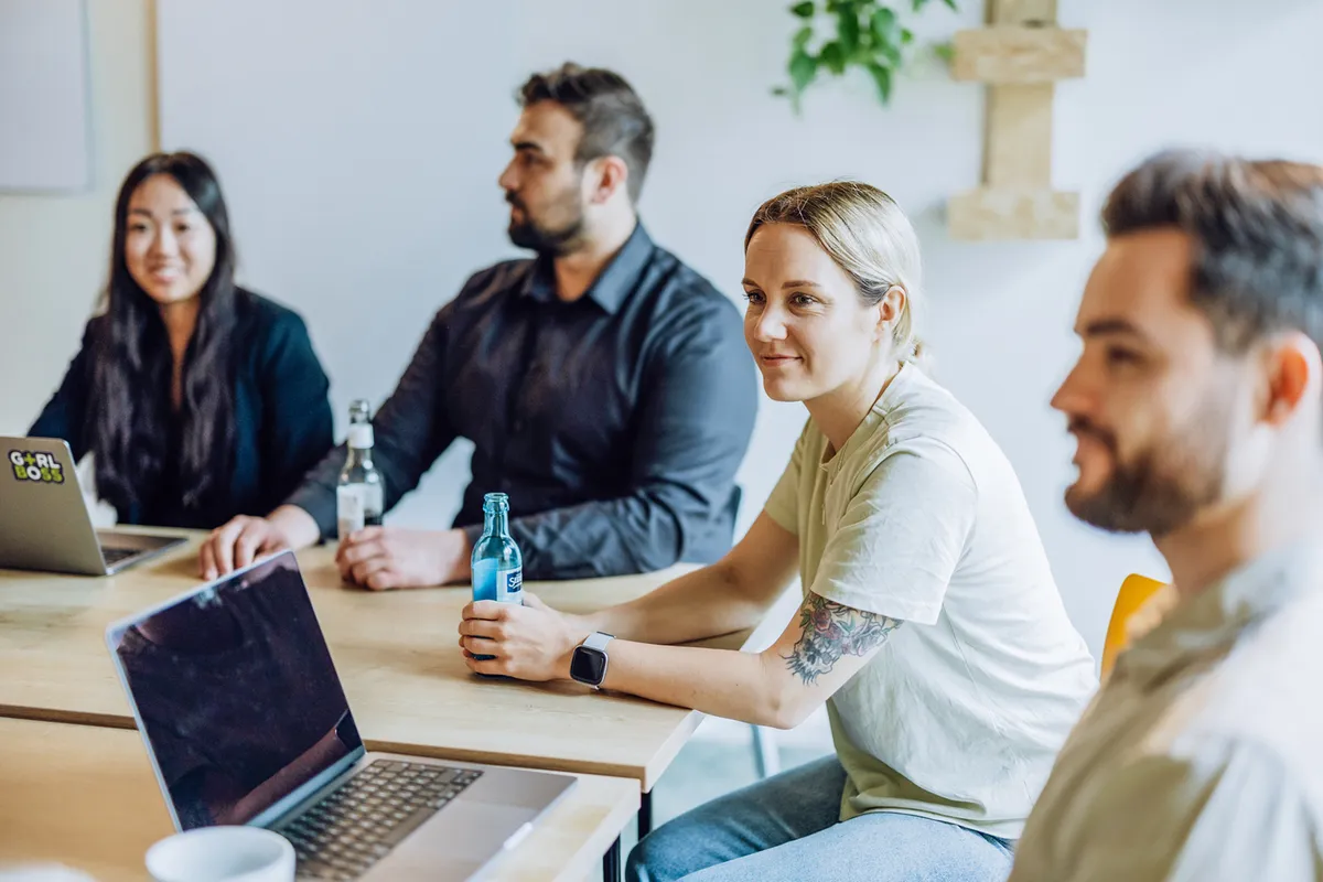 A group of four people sitting around a table with laptops and drinks, engaged in a casual meeting. One person is holding a blue bottle, and another has a laptop with a sticker that reads 'GIRL BOSS'.