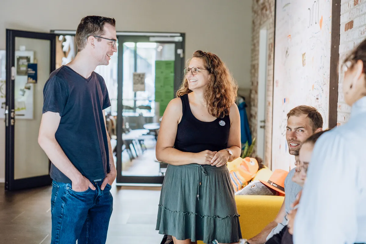 A group of people casually chatting in an office setting, with a man and woman standing and smiling at each other.