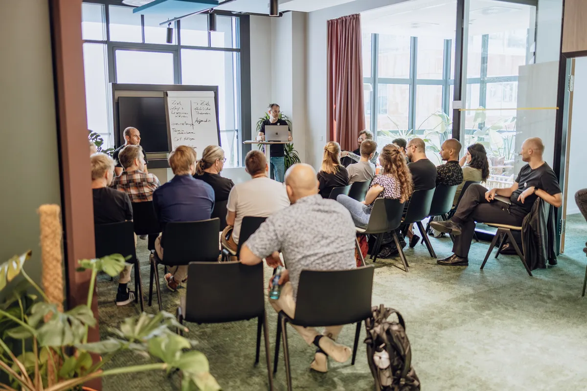 A group of people seated in a conference room, attentively listening to a speaker standing at the front with a laptop and a whiteboard displaying notes.