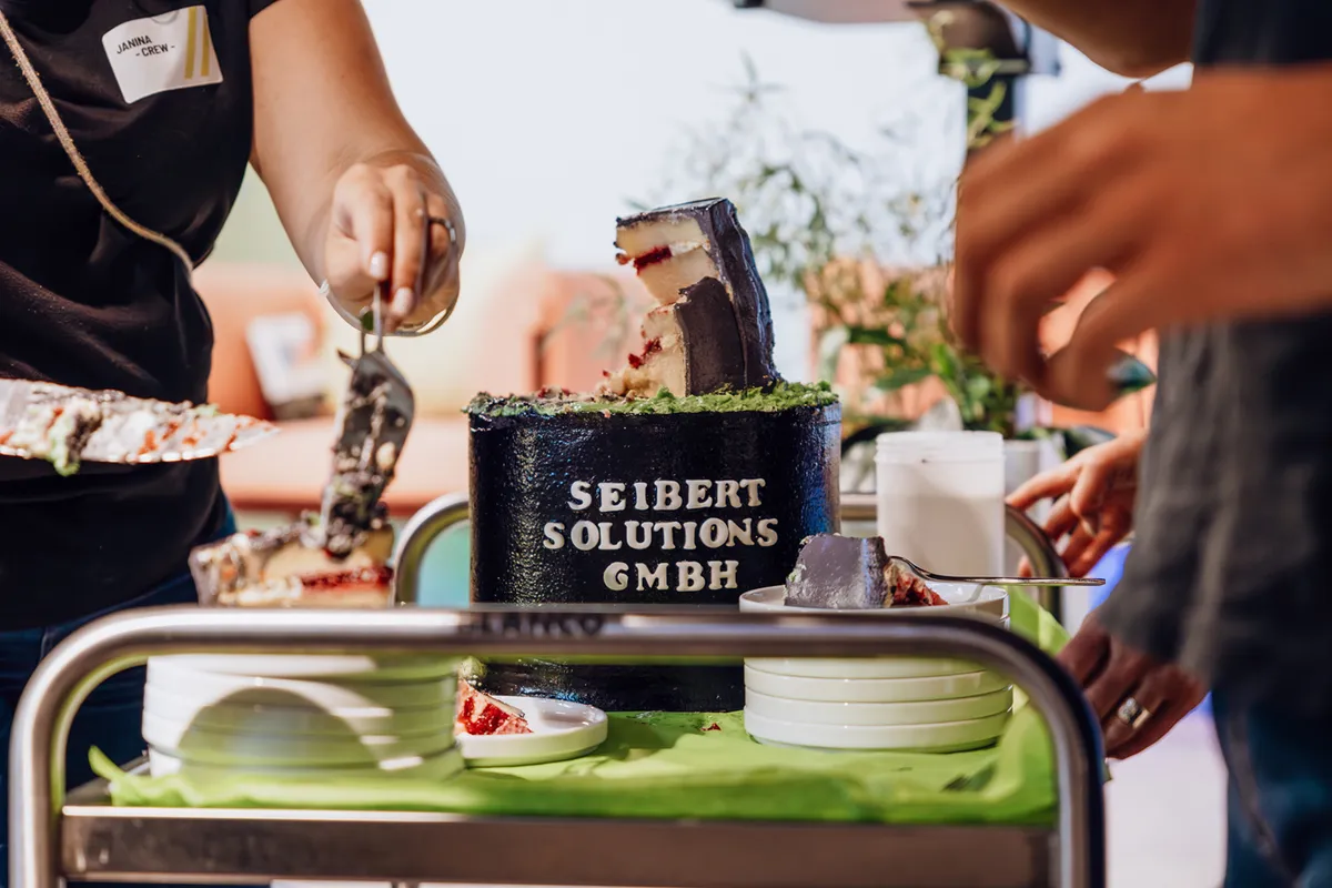 A person cutting a cake with the text 'Seibert Solutions GmbH' on it, placed on a table with plates and utensils.