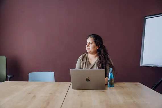 Person at a laptop in a meeting room, looking away in thought