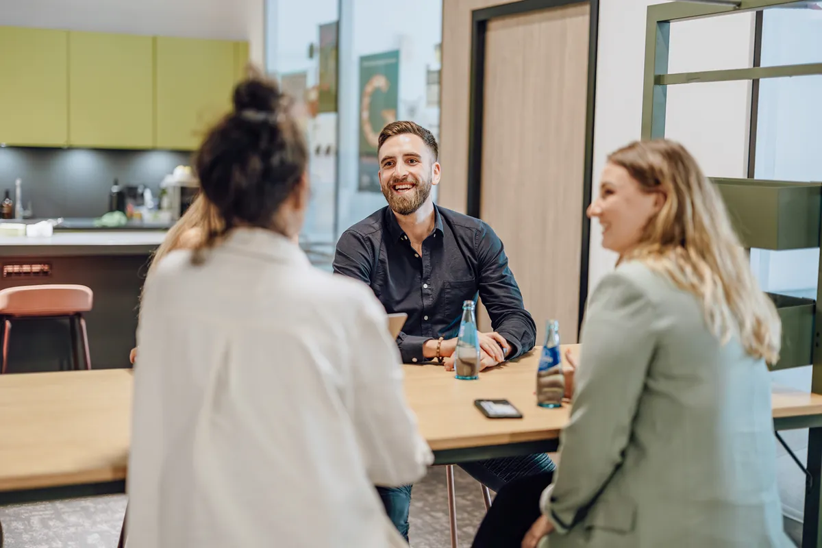 A group of three people sitting around a table in an office setting, engaged in a friendly conversation. They have bottled drinks on the table, and one person is smiling broadly. The background shows a modern office kitchen area.