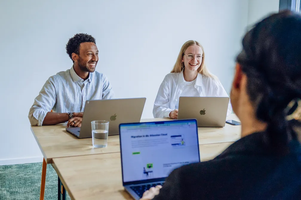 Three people sitting at a table with laptops, engaged in a friendly discussion. Two are facing the camera, smiling, while the third person is seen from behind.