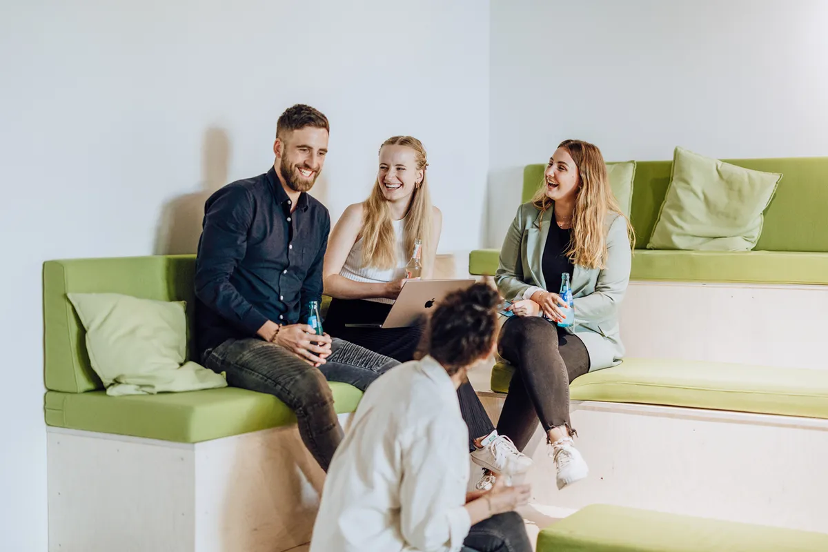 A group of four people sitting on green cushioned benches, smiling and holding drinks, with a laptop open in front of them.