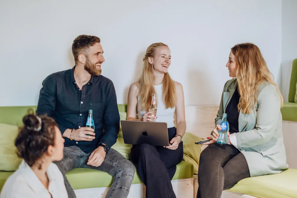 A group of four people sitting on green couches, smiling and holding bottled drinks, with a laptop on one person's lap.