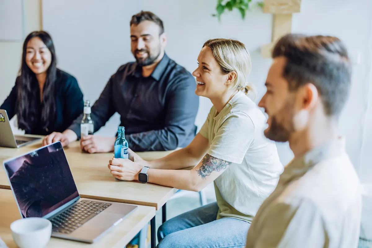 A group of four people sitting around a table with laptops and drinks, smiling and engaged in conversation.