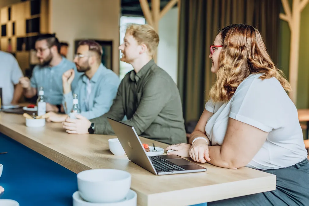 A group of people sitting at a wooden table with laptops and drinks, engaged in a discussion.