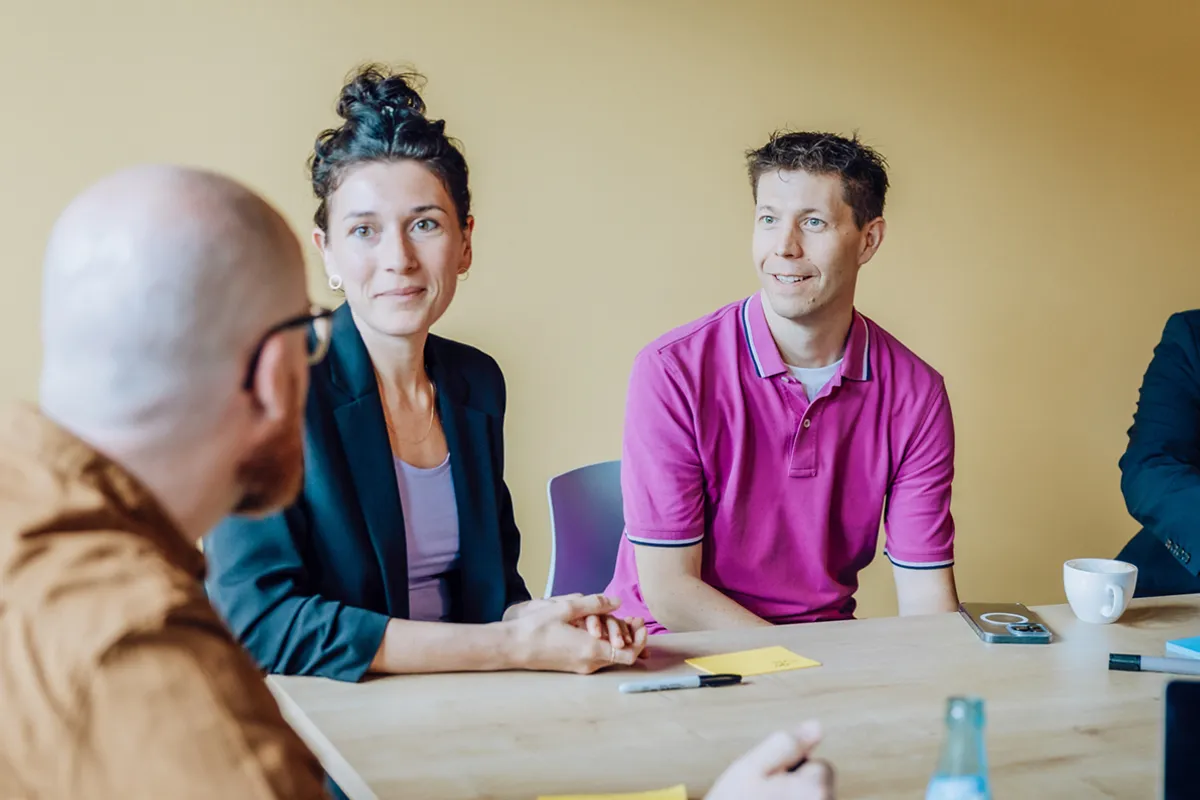 A group of people sitting around a table having a discussion. One person is wearing a pink shirt, and another is wearing a black blazer. There are sticky notes and a marker on the table.
