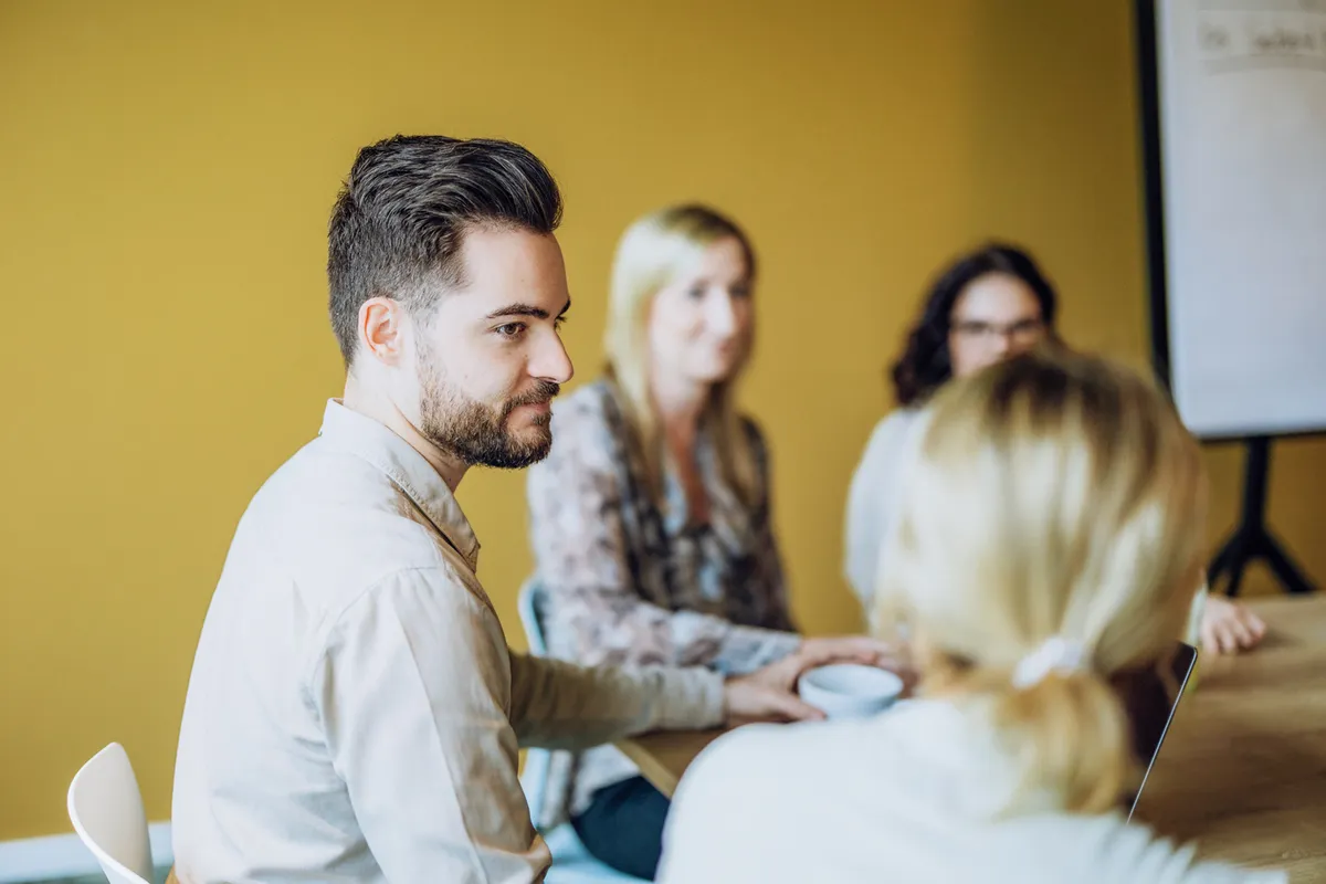 A group of people sitting around a table in a meeting room with a yellow wall. A man in focus is holding a cup, while others are slightly blurred in the background. A flipchart is visible to the side.