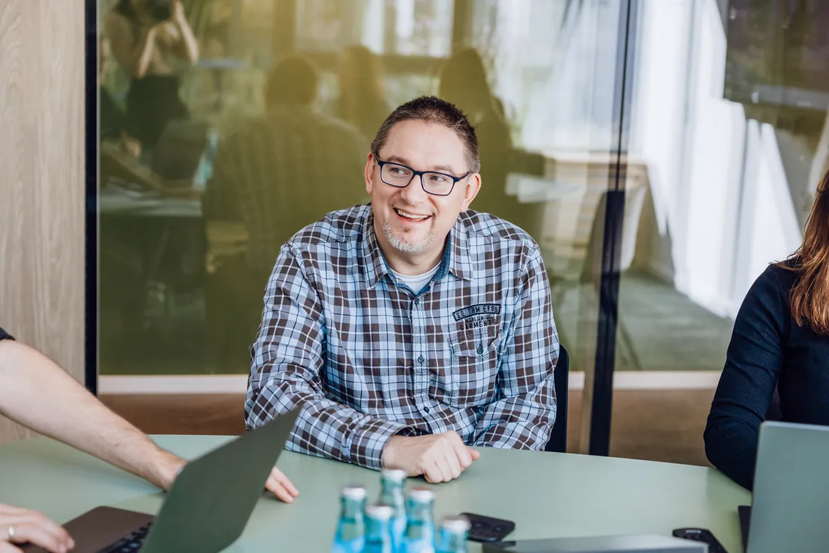 A man in glasses and a plaid shirt smiling while sitting at a table with laptops and water bottles.