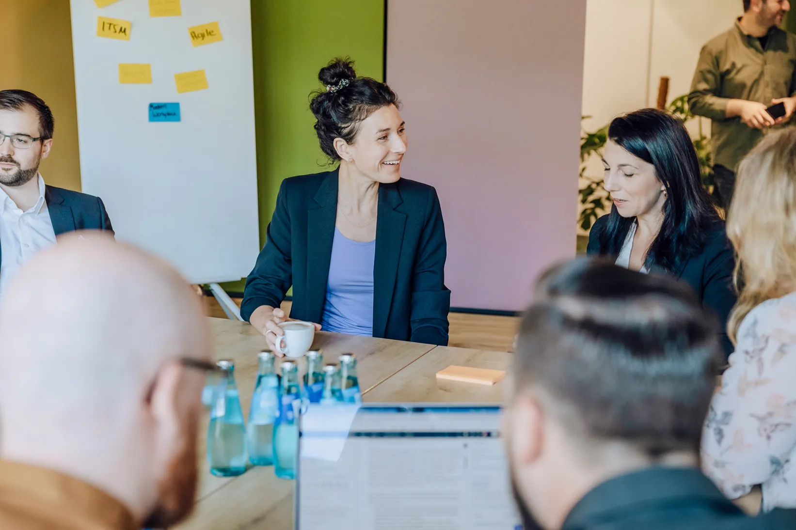 Group of people in a meeting room with a flipchart and sticky notes, discussing around a table with water bottles and a laptop.