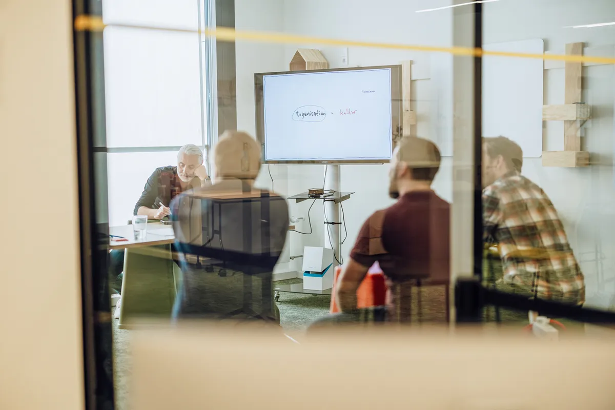 A group of people in a meeting room viewed through a glass wall, with a presentation screen displaying text.