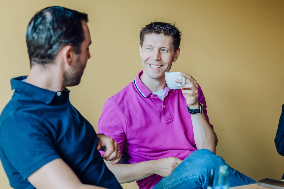 Two men sitting and smiling while having a conversation, one holding a coffee cup, against a yellow background.