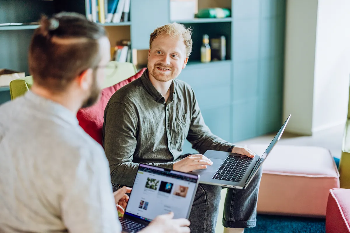 Two men sitting on couches with laptops, engaged in a conversation in a cozy office setting.