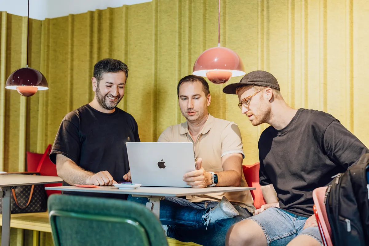 Three men sitting at a table, looking at a laptop screen, with a yellow background and red pendant lights.