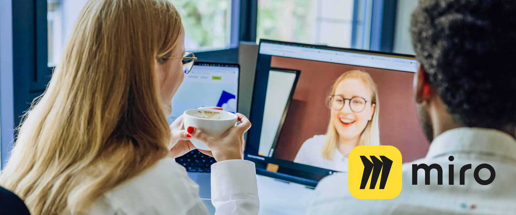 two employees drinking a coffee and holding a virtual meeting on Google Meet with the Miro logo in the foreground