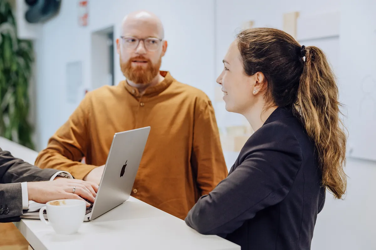 Two people having a conversation at a high table with a laptop and a cup of coffee, in a modern office setting.