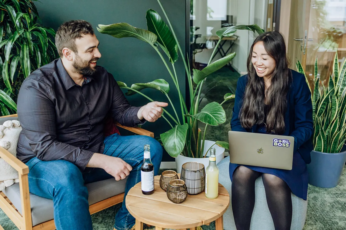Two people sitting in a modern office space with plants, smiling and talking. One person is holding a laptop with a sticker, and there are drinks on a small table between them.