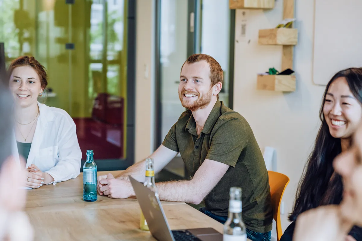A group of people sitting around a table in an office setting, smiling and engaged in conversation. Bottles and a laptop are on the table.