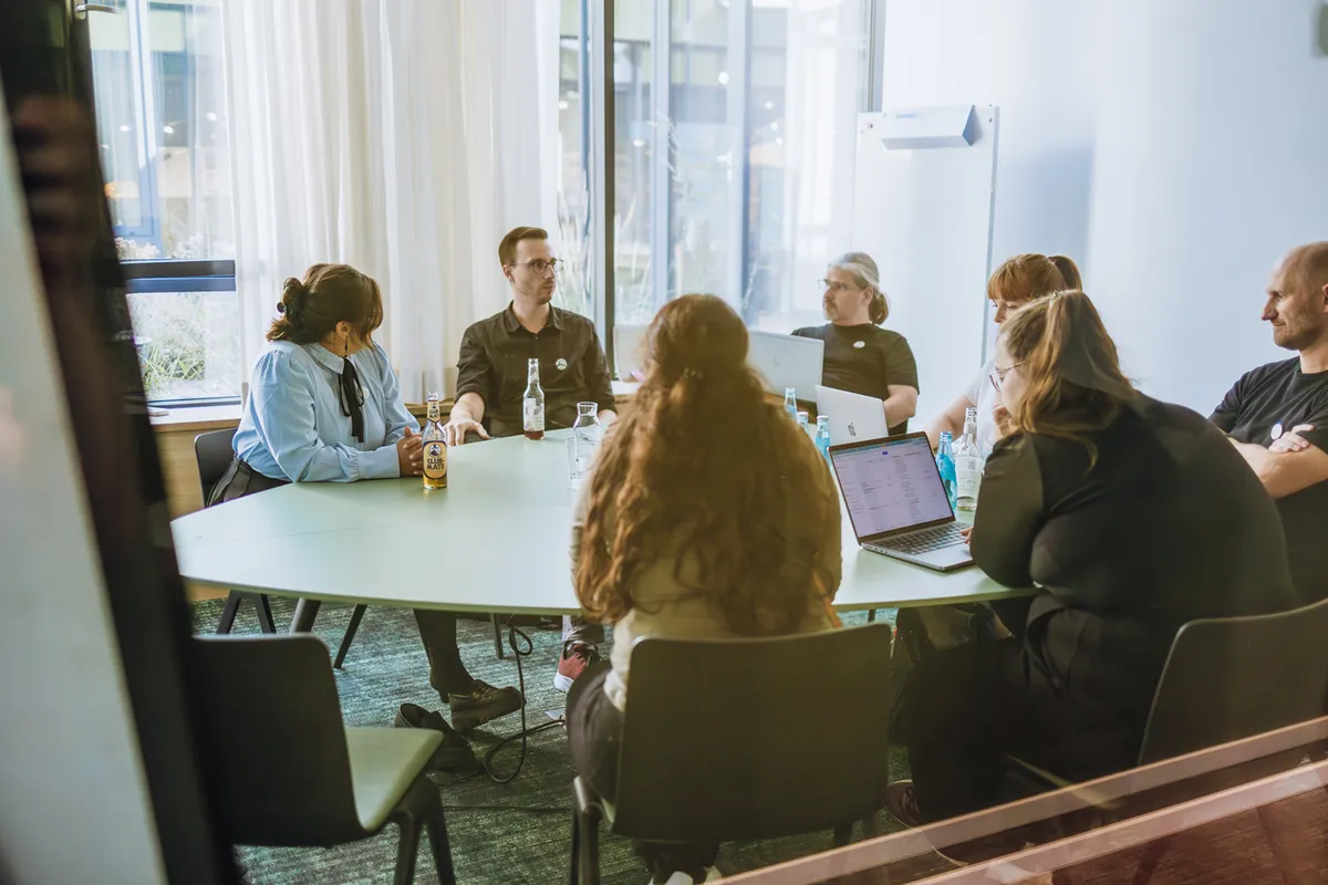 A group of people sitting around a table in a meeting room, with laptops and bottles on the table. They are engaged in discussion, with a whiteboard in the background and large windows letting in natural light.