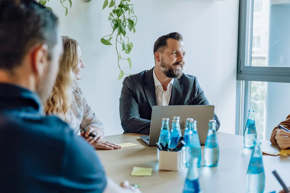 A group of people sitting around a table with laptops and bottled water, engaged in a meeting. A man in a suit is smiling and talking, with plants hanging in the background.