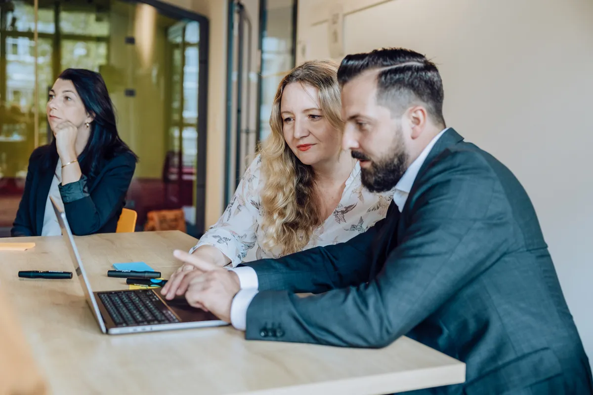 Three people in a meeting room, two focused on a laptop and one listening attentively.