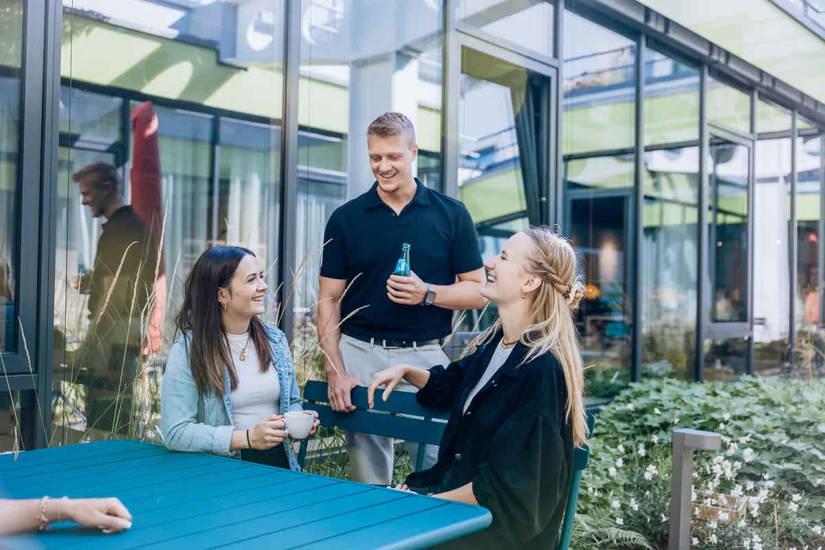 Three people enjoying a conversation outdoors at a blue table, surrounded by greenery and glass buildings.