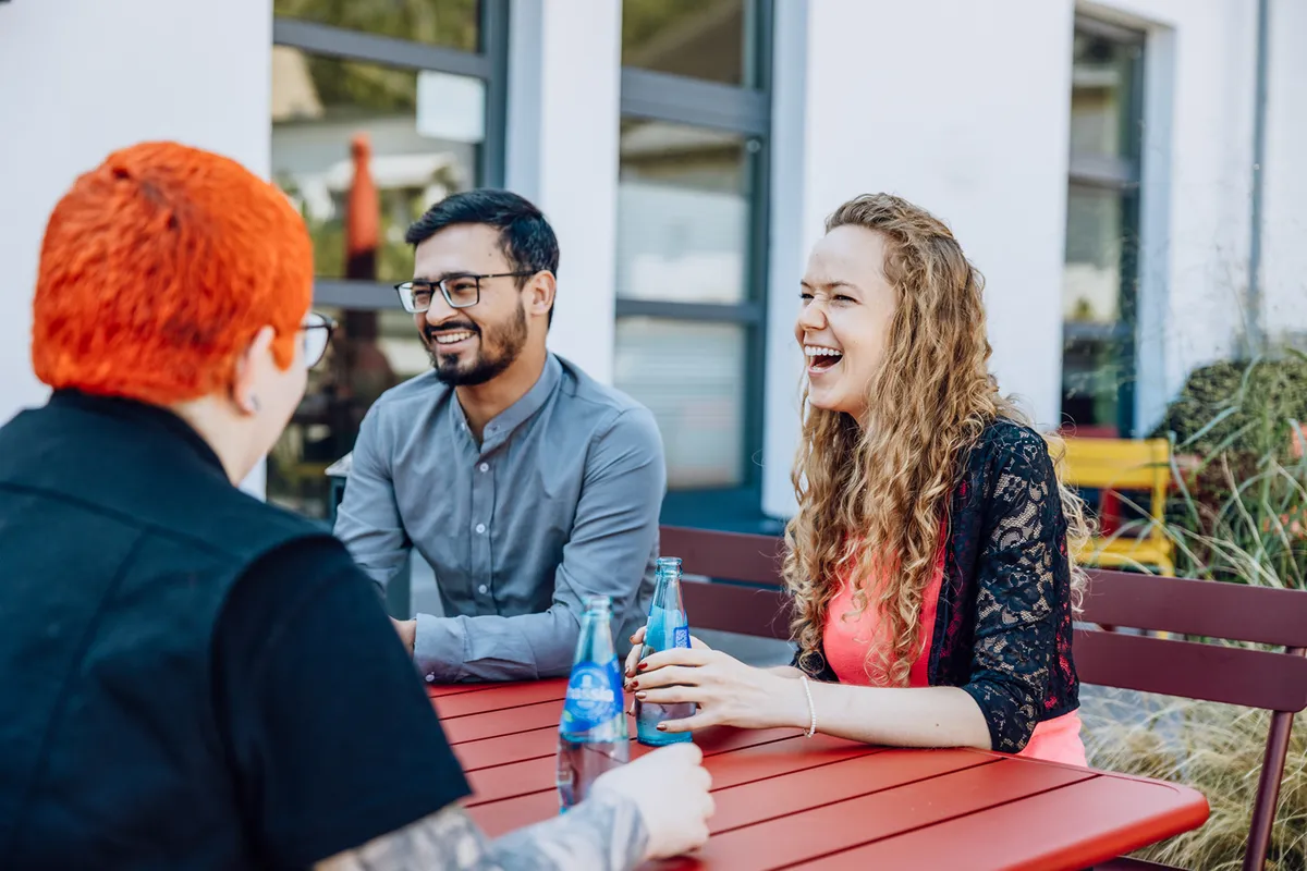 Three people sitting at a red outdoor table, smiling and holding bottled drinks, engaged in a lively conversation.