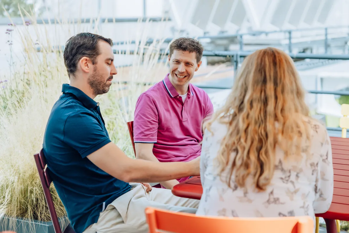 Three people sitting outdoors at a table, engaged in conversation. The setting is bright and airy with plants in the background.