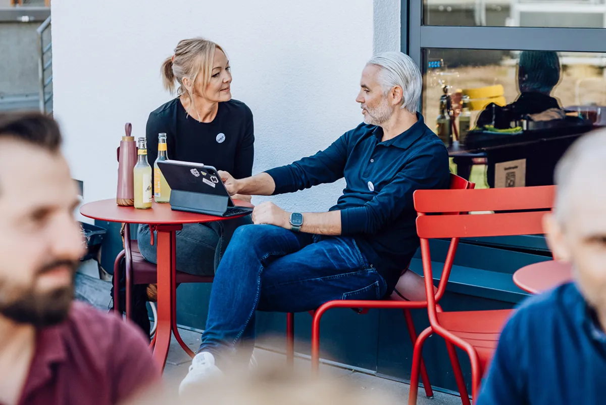 Two people sitting at a red outdoor table, engaged in conversation with a tablet and drinks on the table.