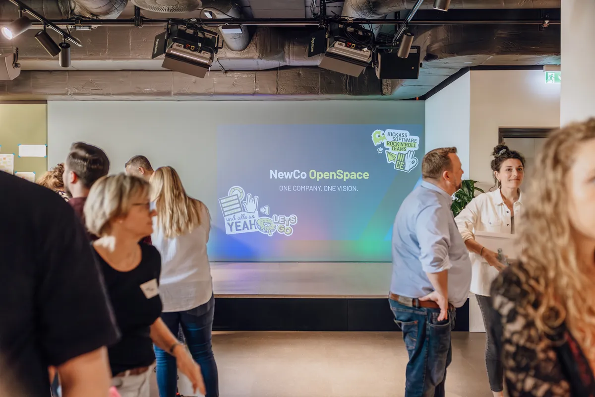 People mingling in front of a presentation screen displaying the text 'NewCo OpenSpace' with colorful graphics.