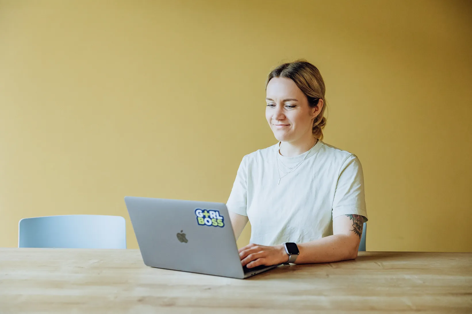 A person sitting at a wooden table using a laptop with a sticker on it, against a yellow wall.