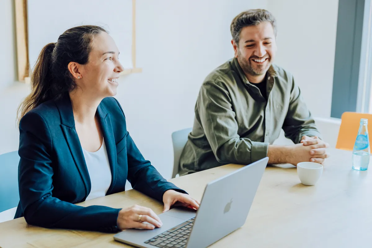 Two people sitting at a table, smiling and engaged in conversation. One person is using a laptop, and there is a cup and a bottle of water on the table.