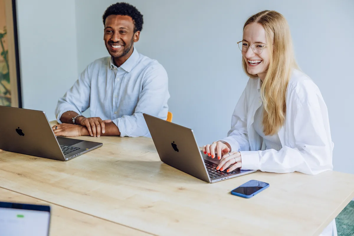 Two people sitting at a table with laptops, smiling and engaged in conversation.