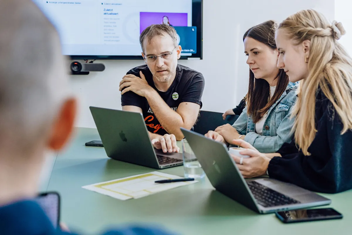 A group of people sitting at a table with laptops, engaged in a discussion. A screen is visible in the background displaying a presentation.