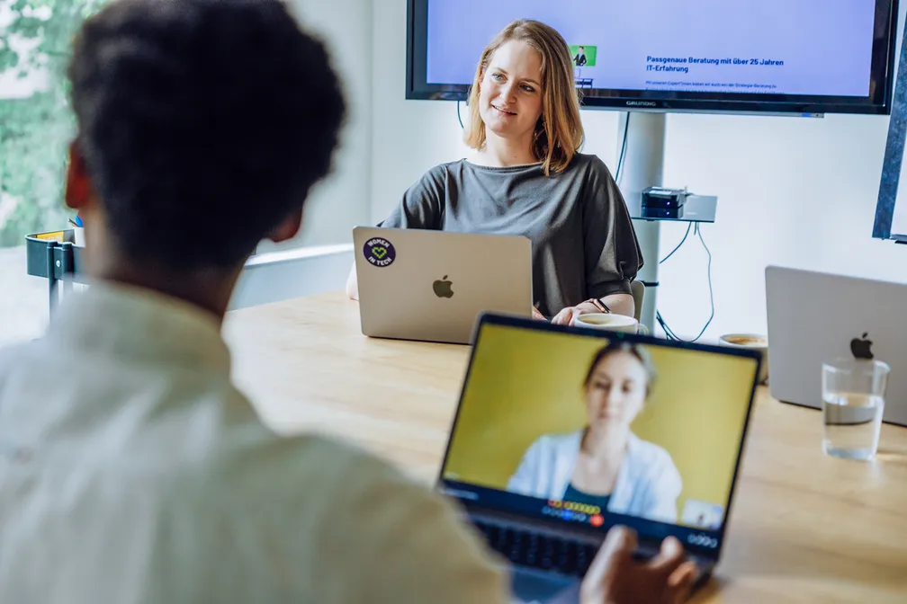 Two people having a video call meeting in an office setting, with laptops and a screen in the background.