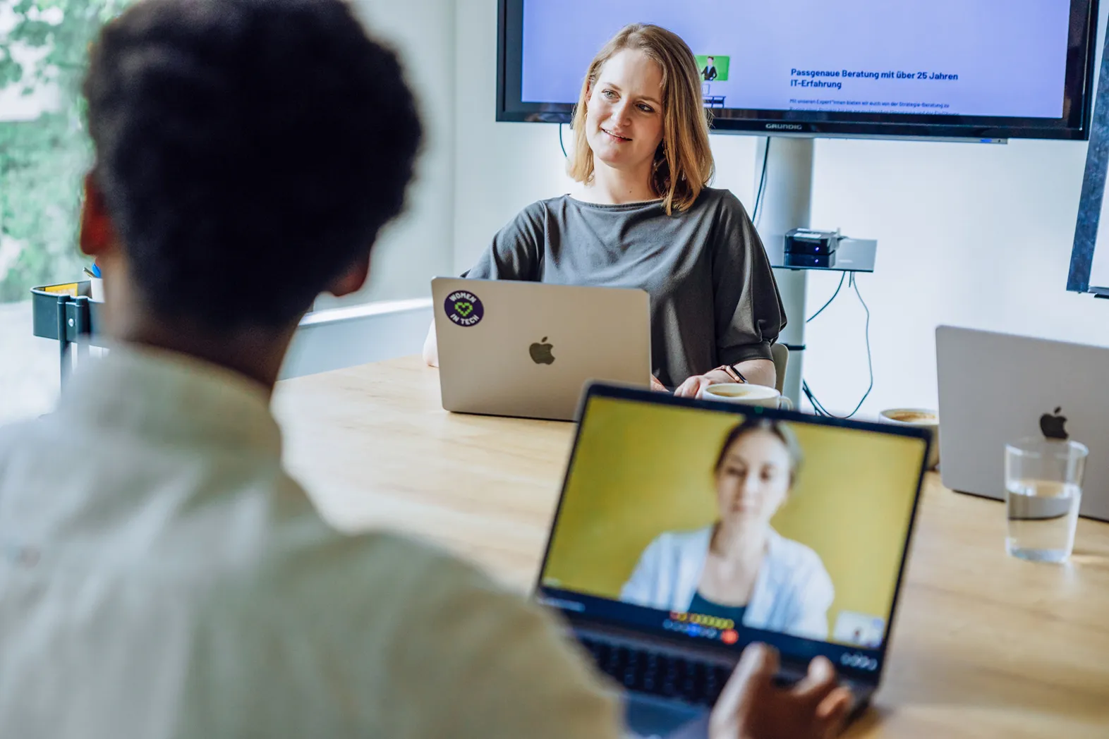 Two people having a video call meeting in an office setting, with laptops and a screen in the background.